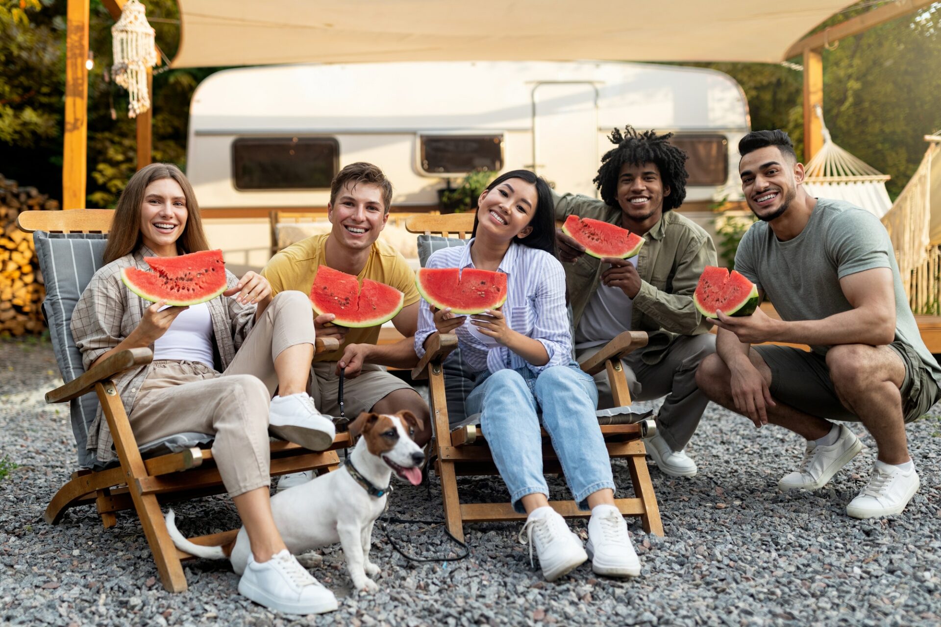 Happy diverse friends eating watermelon near RV, sitting in lounge chairs and enjoying autumn