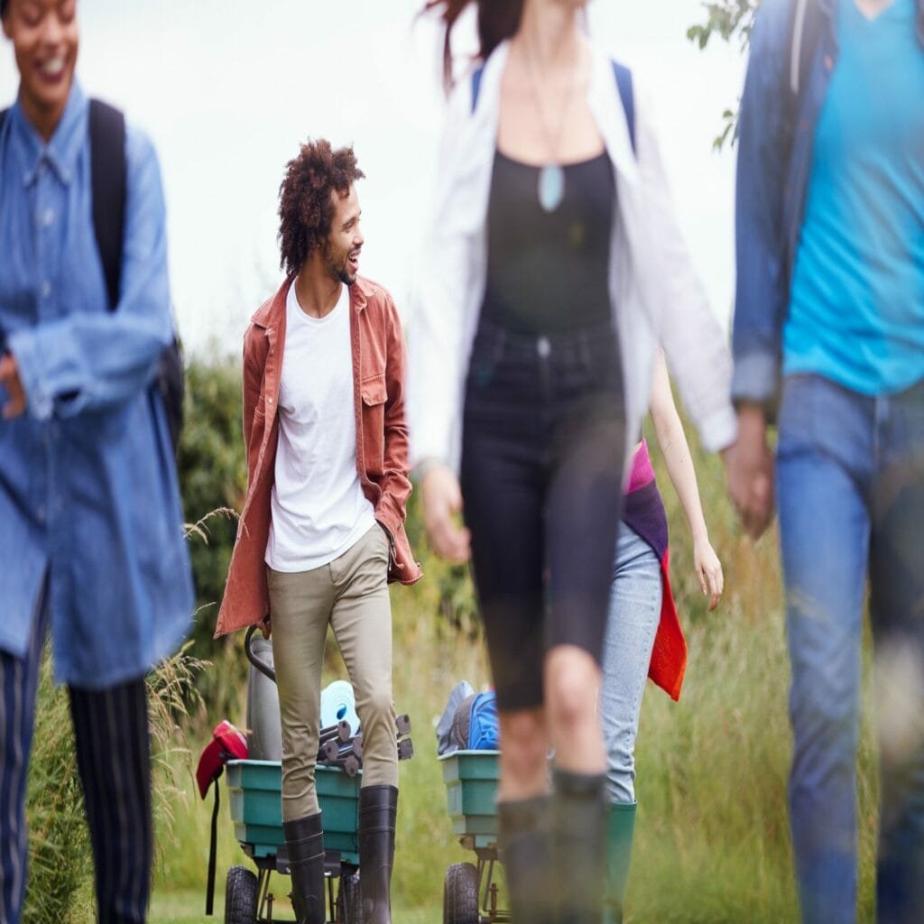 Group Of Excited Young Friends Carrying Camping Equipment Through Field To Music Festival