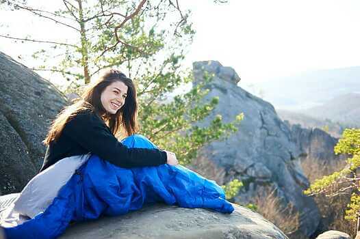 Woman sitting alone in sleeping bag on big mountain rock and smiling