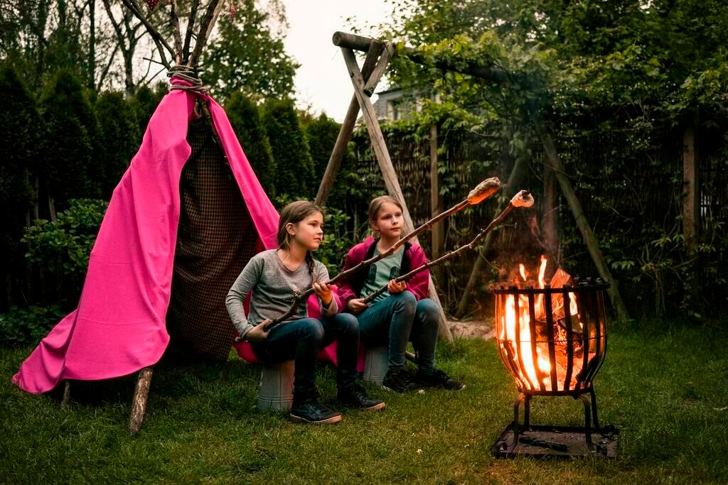 Two girls wearing feather headdress, with twist bread on camp fire