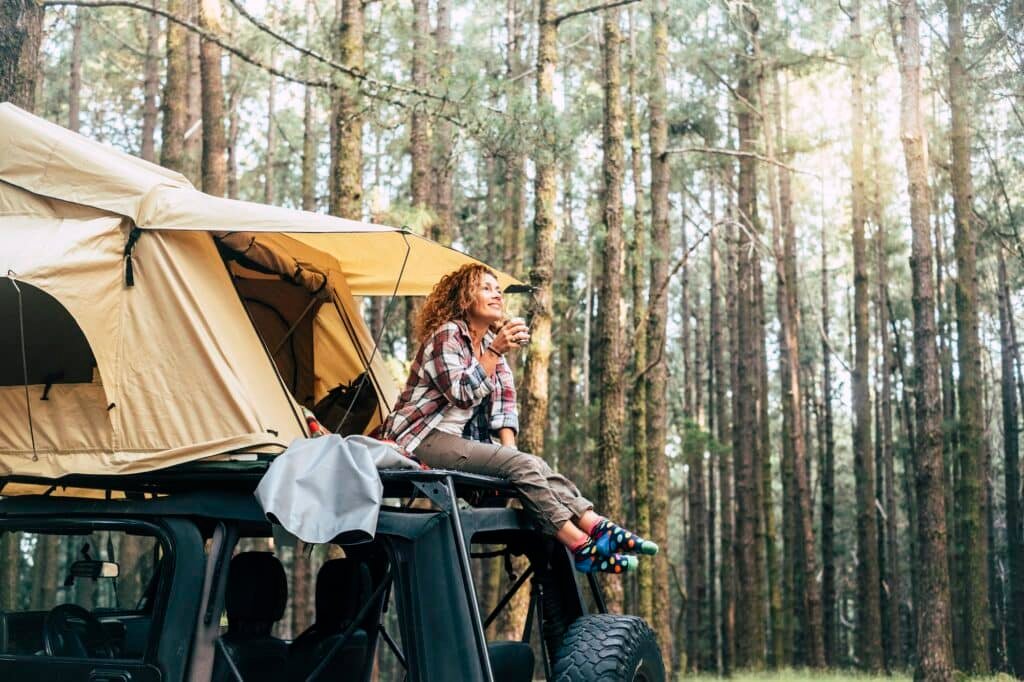 Young woman sitting outside tent on the roof of the car and drinking coffee in forest. Woman relax