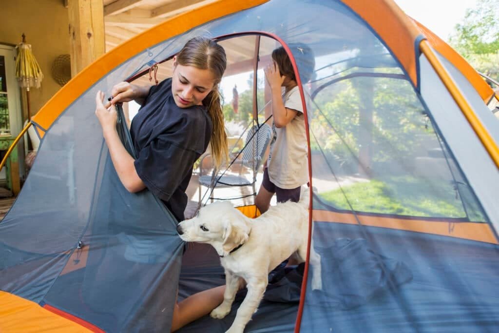Familienfreundlich Unterwegs: Ihr Ultimativer Leitfaden für Familienzelte für den Campingurlaub 4 Teenage girl and her younger brother setting up a tent, a cute puppy tugging the tent fabric.