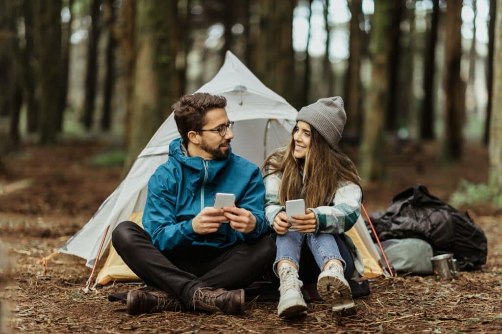 Cheerful young european man and female tourists in jackets resting in forest near tent, typing on