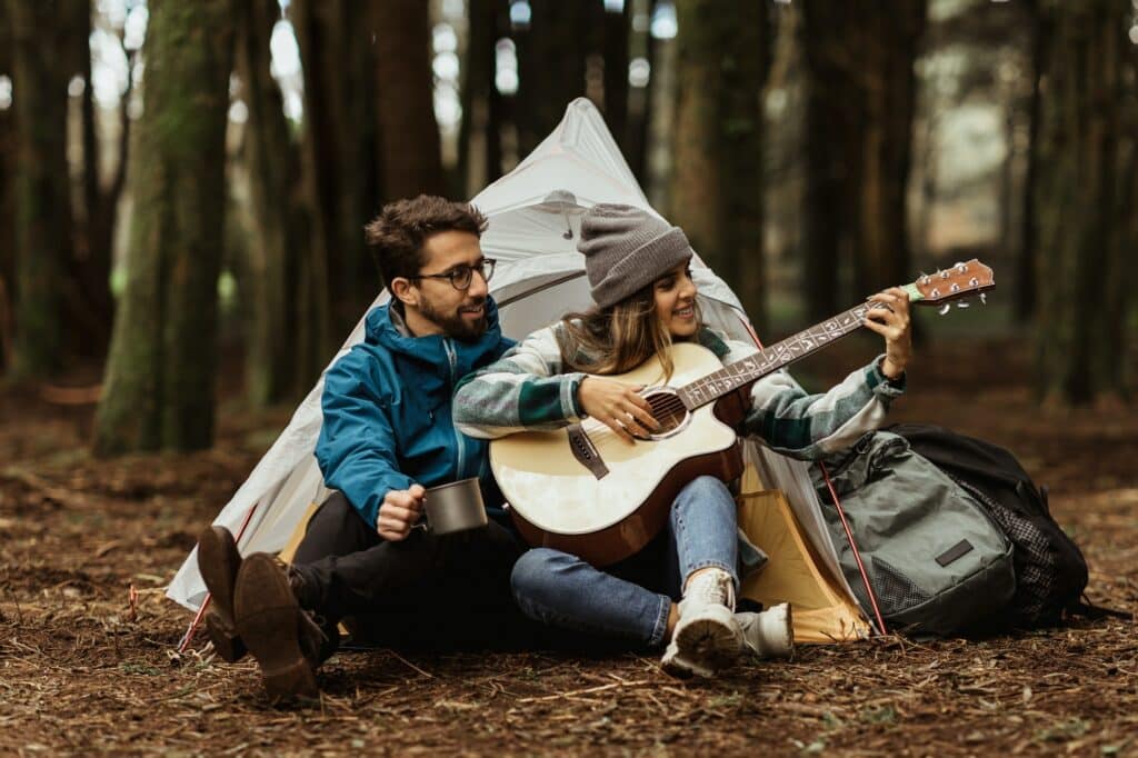 Glad young european male and woman tourists in jackets rest in forest near tent, enjoy cold season