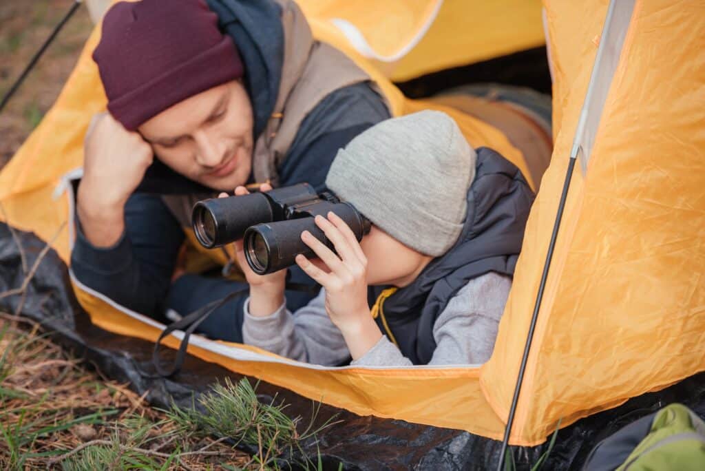 father and son looking through binoculars while lying together in tent
