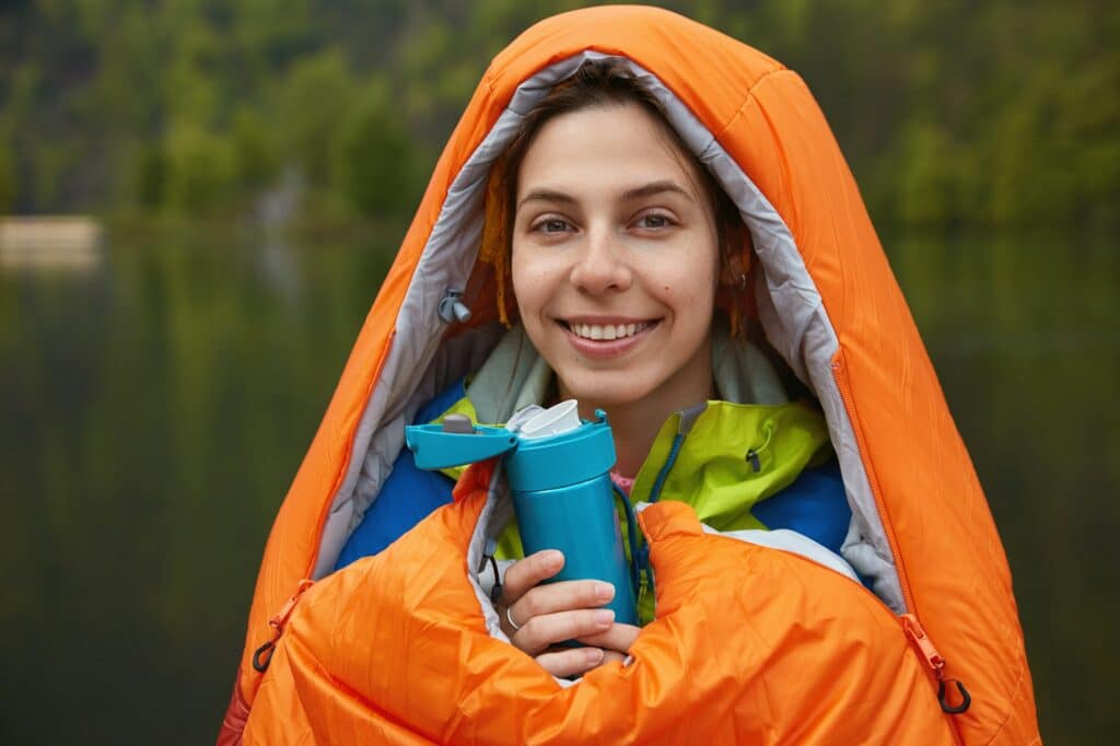 Happy female hiker has camping trip, being cold after rain, warms in sleeping bag, drinks hot tea fr