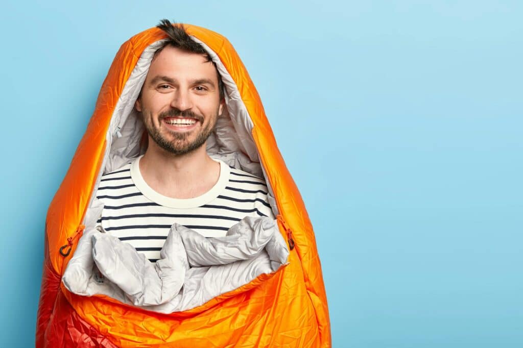 Happy unshaven man vacationist spends holidays near mountains in camping, sleeps in sleeping bag, sm