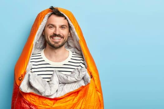 Happy unshaven man vacationist spends holidays near mountains in camping, sleeps in sleeping bag, sm