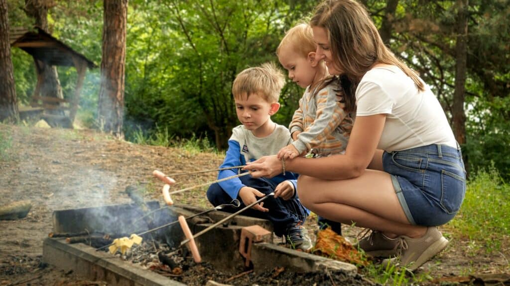 Young mother with two sons cooking sausages on bonfire in camp. Active leisure, children in camping