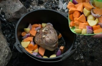 Campfire cooking. Cast iron camp oven with lamb leg inside. Adding chopped vegetables from the bowl