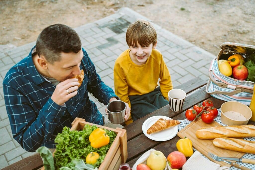 Father dad and school kid boy child eating croissant picnic forest camping site with vegetables