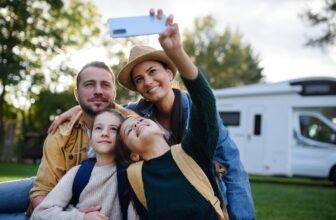 Happy young family with two children taking selfie with caravan at background outdoors.